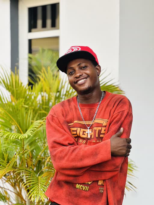 King Triumphant smiling to camera in a red sweatshirt and cap, standing in front of palm fronds.